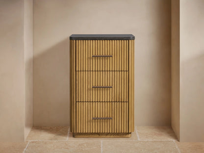 Finnley Short Bath Cabinet: light oak fluted wood, dark stone top, 3 drawers with bronze pulls, on tan wall and stone floor.