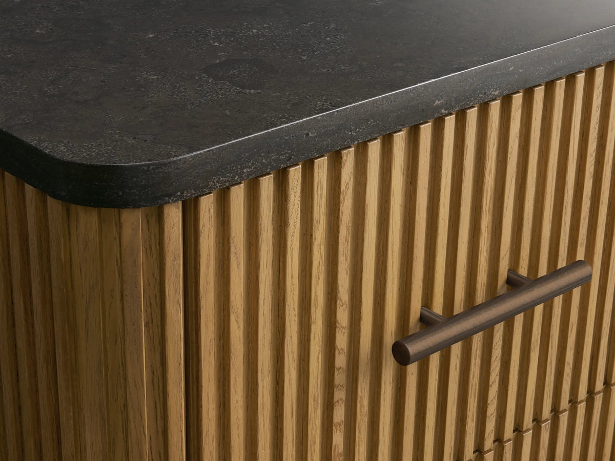 Finnley Short Bath Cabinet: light oak wood with vertical fluting, three drawers, dark bronze handles, dark bluestone top.