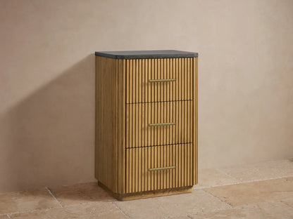 Finnley Short Etagere with fluted oak wood design, black stone top, and three drawers with brass handles, set in neutral room.