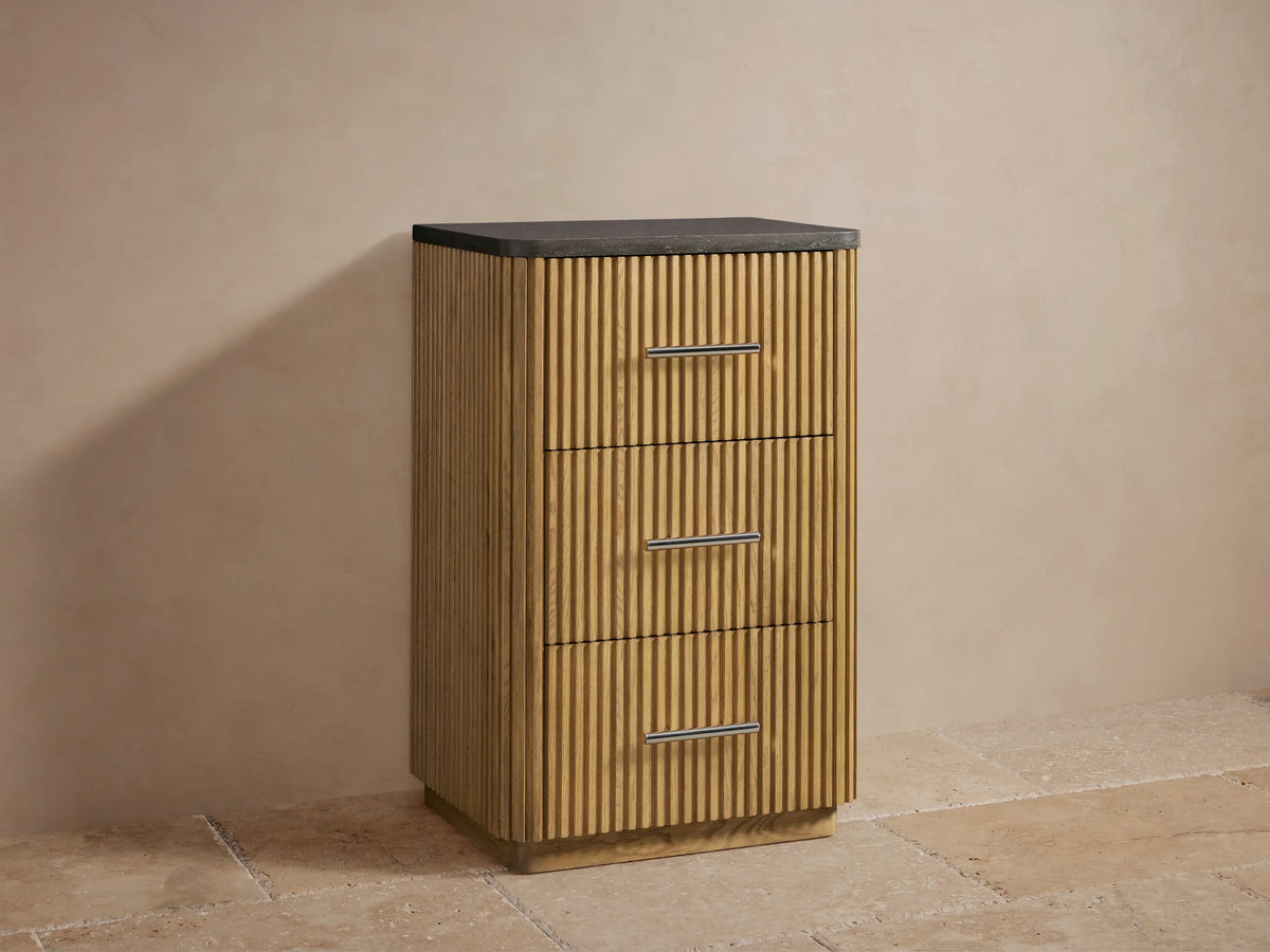 Finnley Short Etagere featuring vertical oak slats, black stone top, three drawers with polished nickel handles, in a beige alcove.