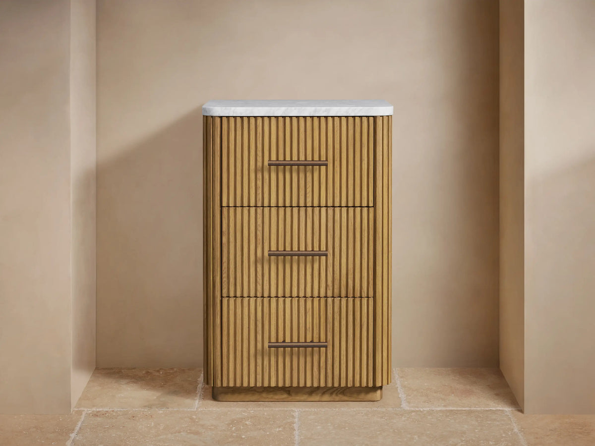 Finnley Short Bath Cabinet: light oak reeded wood cabinet with white marble top, three drawers with bronze pull handles, in a beige bathroom.