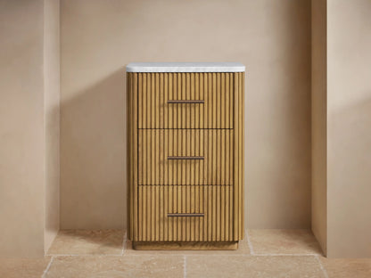 Finnley Short Bath Cabinet: light oak reeded wood cabinet with white marble top, three drawers with bronze pull handles, in a beige bathroom.