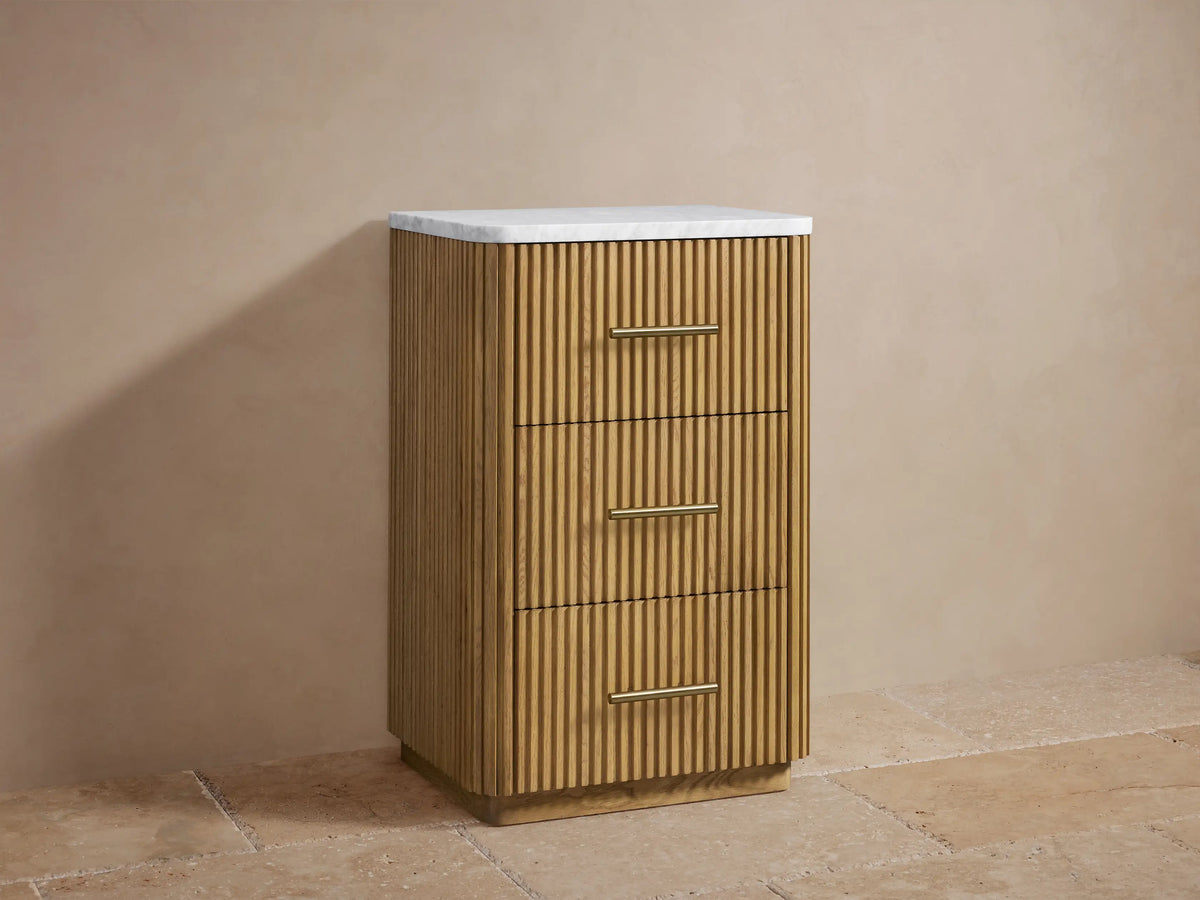 Finnley Short Etagere with vertical slat oak wood design, three drawers, brass handles, and a sealed stone top, against a neutral background.