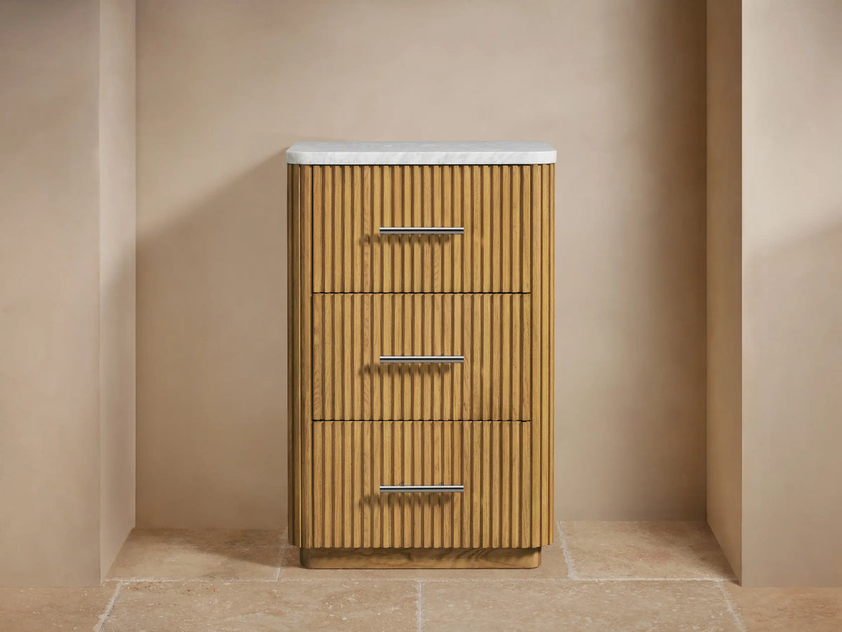 Finnley Short Etagere with a stone top, vertical slatted wood pattern, three drawers with metallic handles, and decorative items on top.