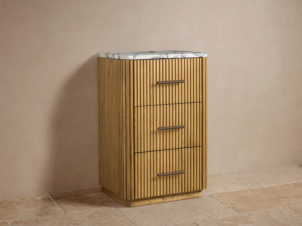 Finnley Short Bath Cabinet, natural oak wood with vertical fluting, white marble top. Three drawers with bronze handles. Towels & decor on top.