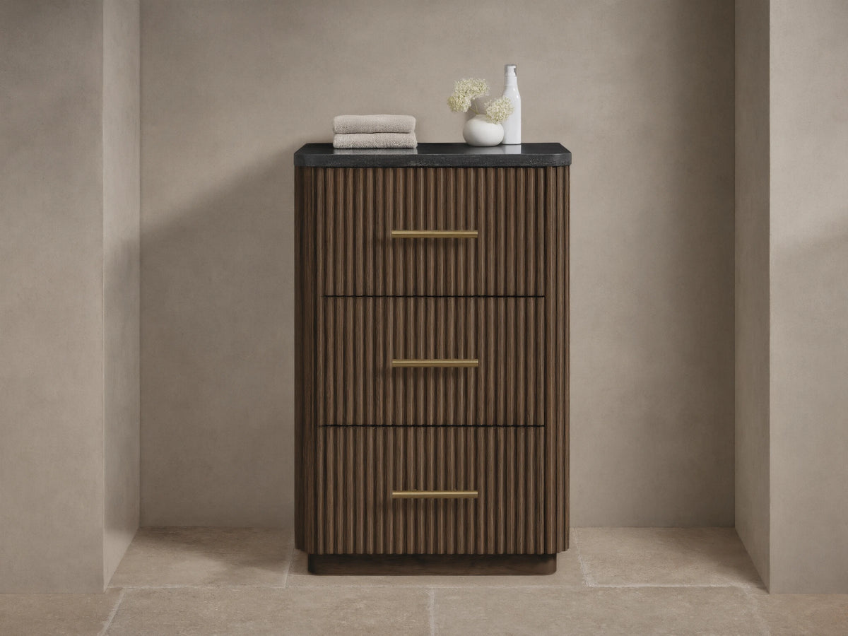 Finnley Short Bath Cabinet. Dark brown fluted wood, 3 drawers with brass pulls, black stone top. White towels and vase on top.
