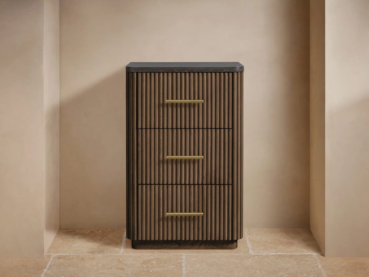 Finnley Short Bath Cabinet. Dark brown fluted wood, 3 drawers with brass pulls, black stone top. White towels and vase on top.