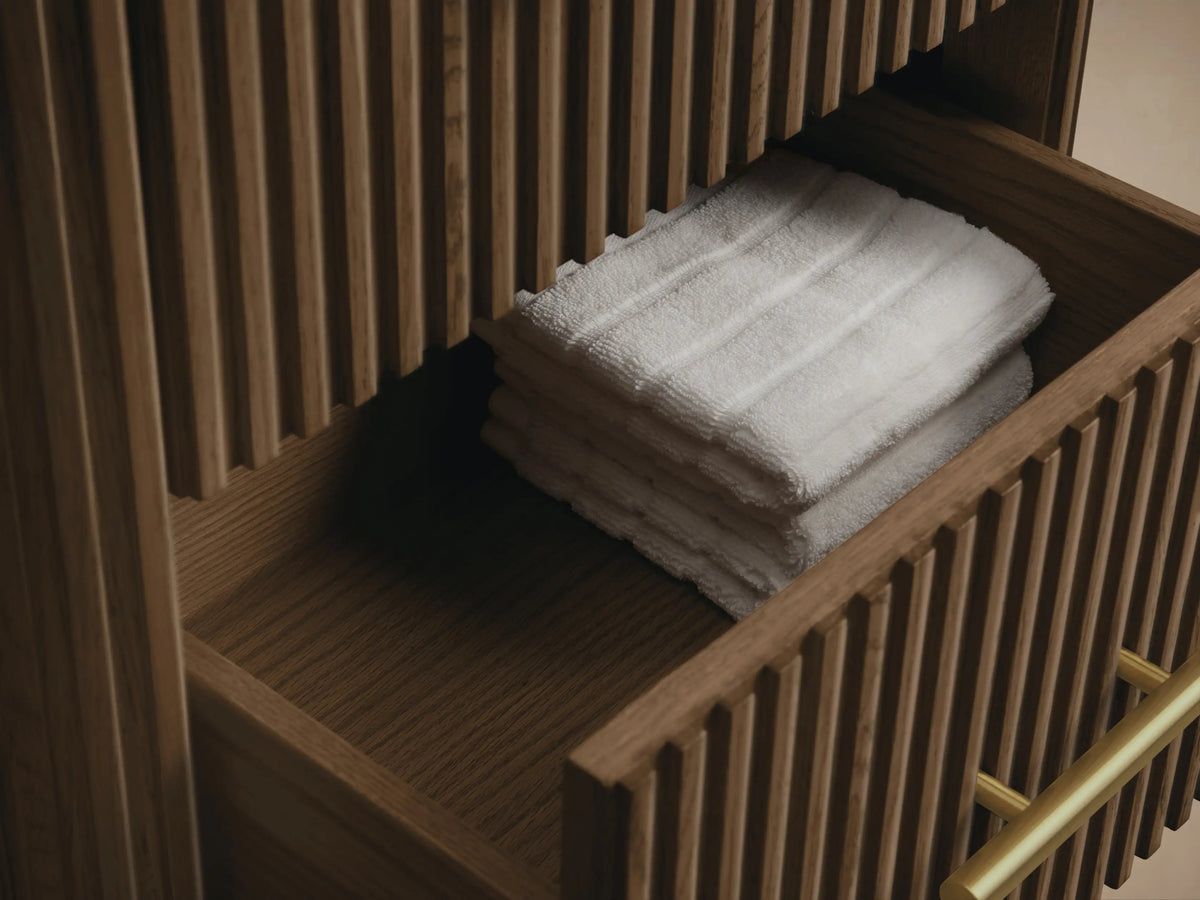 Finnley Short Bath Cabinet, dark fluted oak wood with a dark stone top & 3 horizontal brass pulls, in a warm beige room.