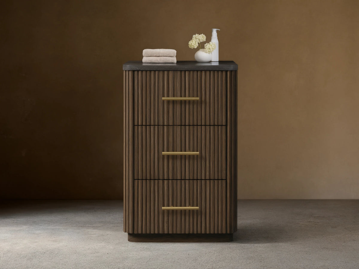 Finnley Short Bath Cabinet. Dark brown fluted wood, 3 drawers with brass pulls, black stone top. White towels and vase on top.