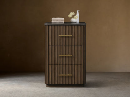 Finnley Short Bath Cabinet. Dark brown fluted wood, 3 drawers with brass pulls, black stone top. White towels and vase on top.