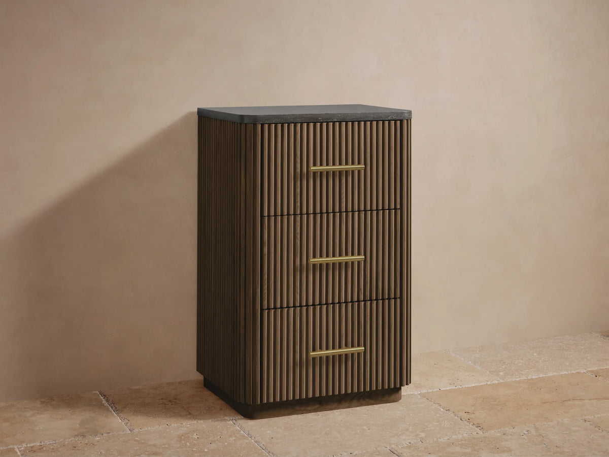 Finnley Short Bath Cabinet with dark, textured stone top, reeded oak wood, and a horizontal brass handle. Close-up, angled.
