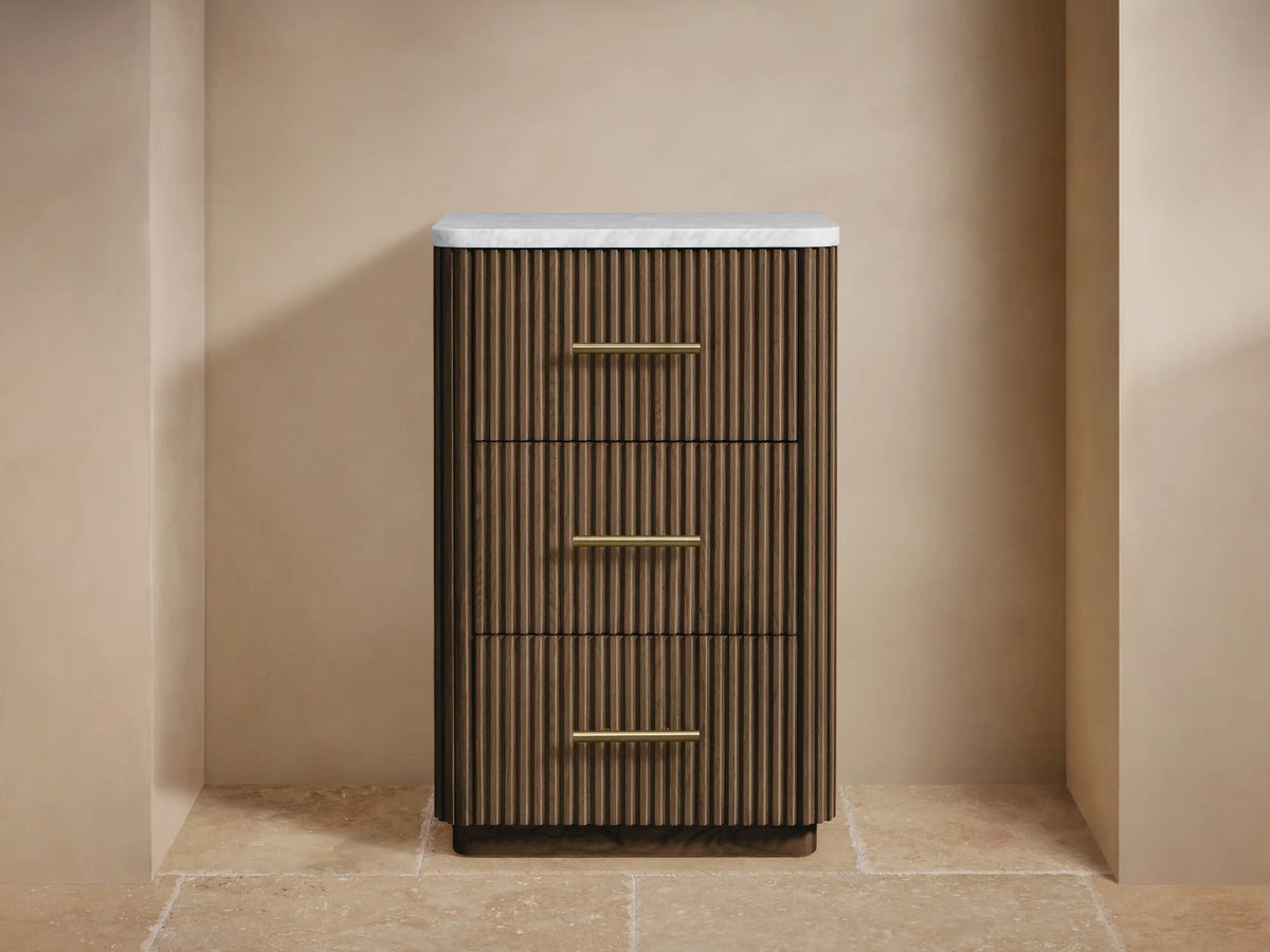 Finnley Short Bath Cabinet. Dark oak fluted wood cabinet with white Carrara marble top, 3 drawers, brass pulls. Towels & flowers on top.