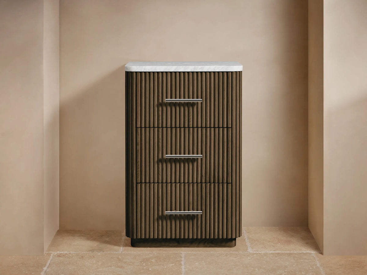 Finnley Short Bath Cabinet: dark fluted wood, 3 drawers with silver pulls, white marble top, towels & flowers on top.