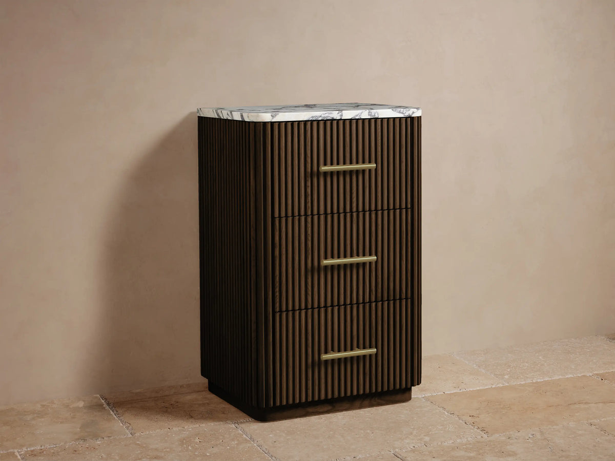 Finnley Short Bath Cabinet: Dark brown slatted wood with 3 drawers, brass pulls, and a white marble top, against a beige wall.