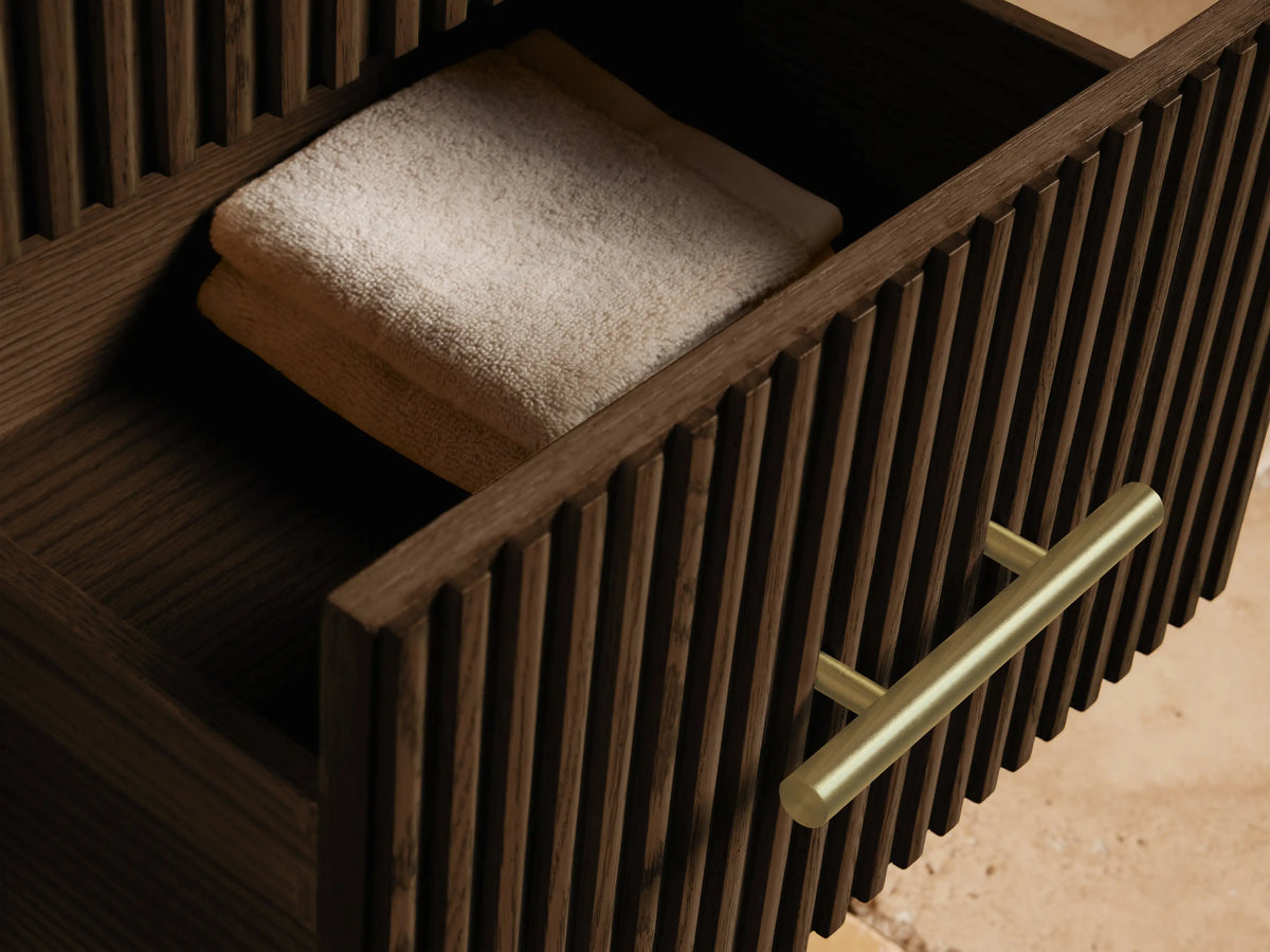 Close-up of Finnley Short Bath Cabinet. Light marble top with dark veins over dark fluted oak body and brass handle.
