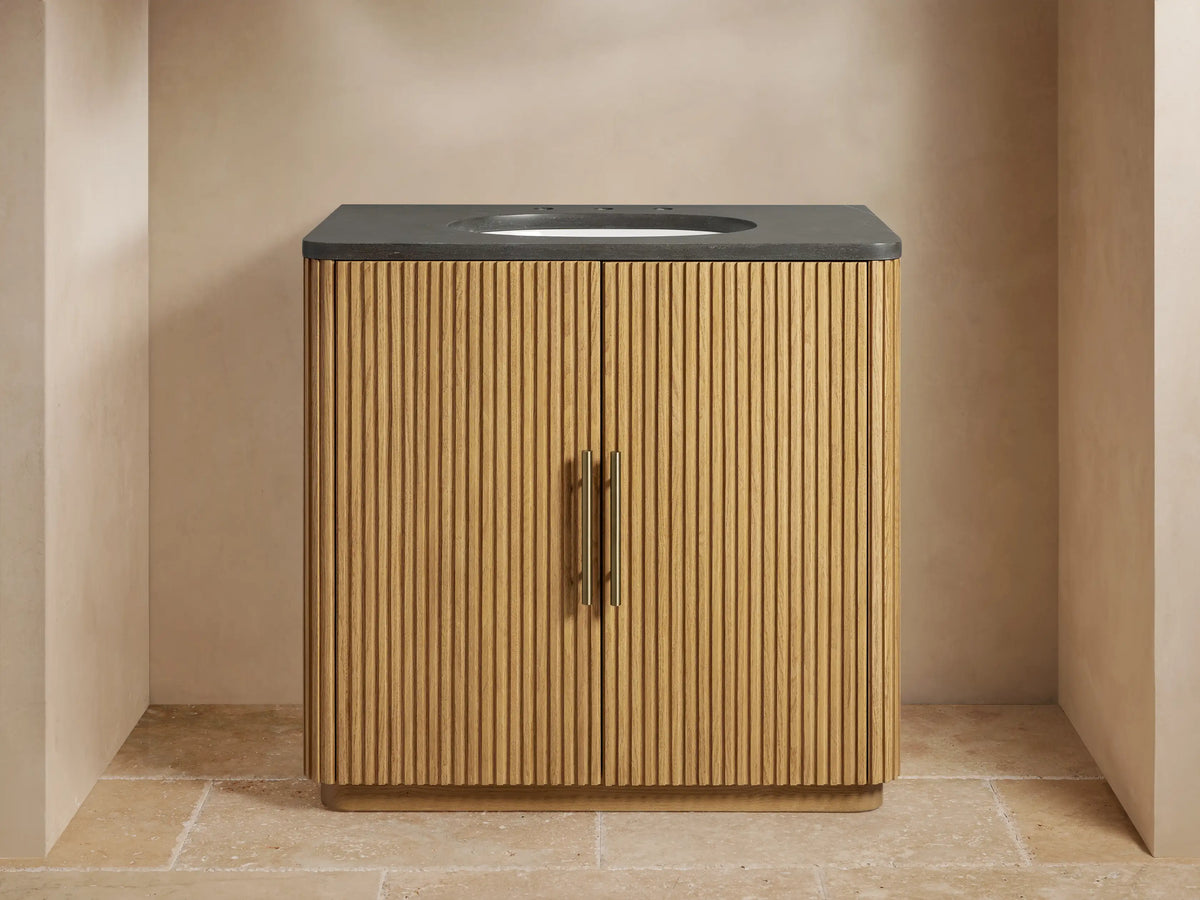 Open Finnley Single Vanity cabinet with ribbed oak wood door. Shelves inside hold folded towels and a wooden tray with a brush.