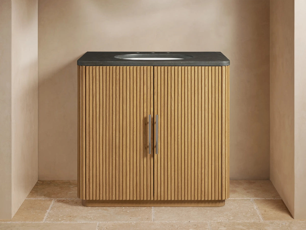 Finnley Single Vanity with vertical ribbed oak wood design, black stone top, oval sink, and metal handles, against a beige wall.