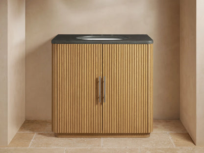 Finnley Single Vanity with vertical ribbed oak wood design, black stone top, oval sink, and metal handles, against a beige wall.