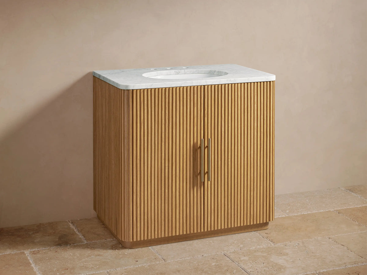 Finnley Single Vanity with oak wood slatted facade, marble top, oval sink, and brass faucets against beige wall.