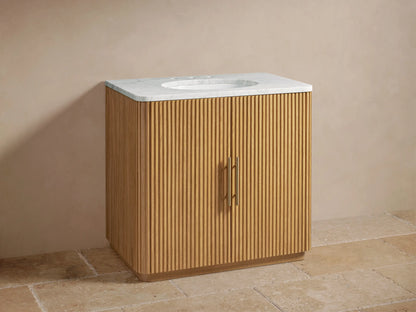 Finnley Single Vanity with oak wood slatted facade, marble top, oval sink, and brass faucets against beige wall.