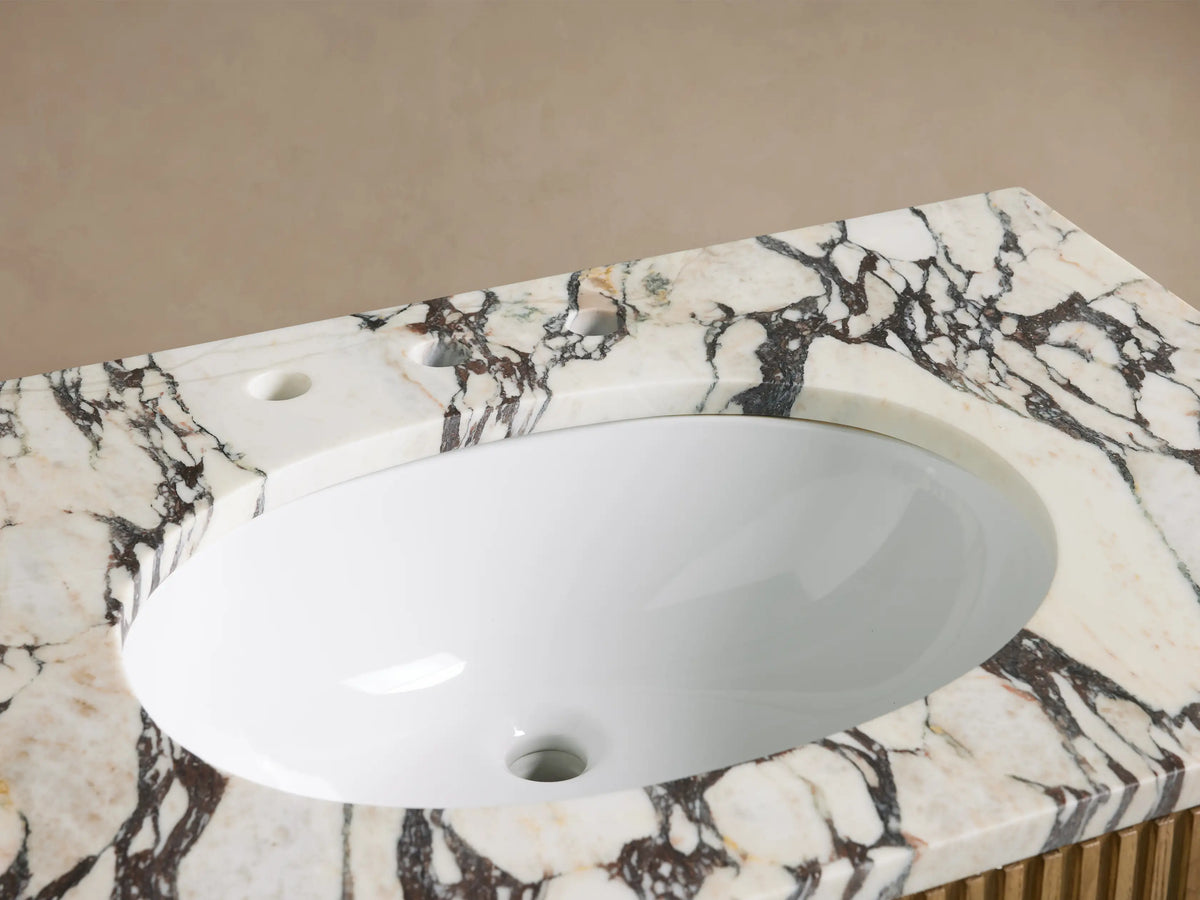 Finnley Single Vanity: Close-up of white oval sink in honed marble top with dark veining, widespread bronze faucet, stone soap dispenser.