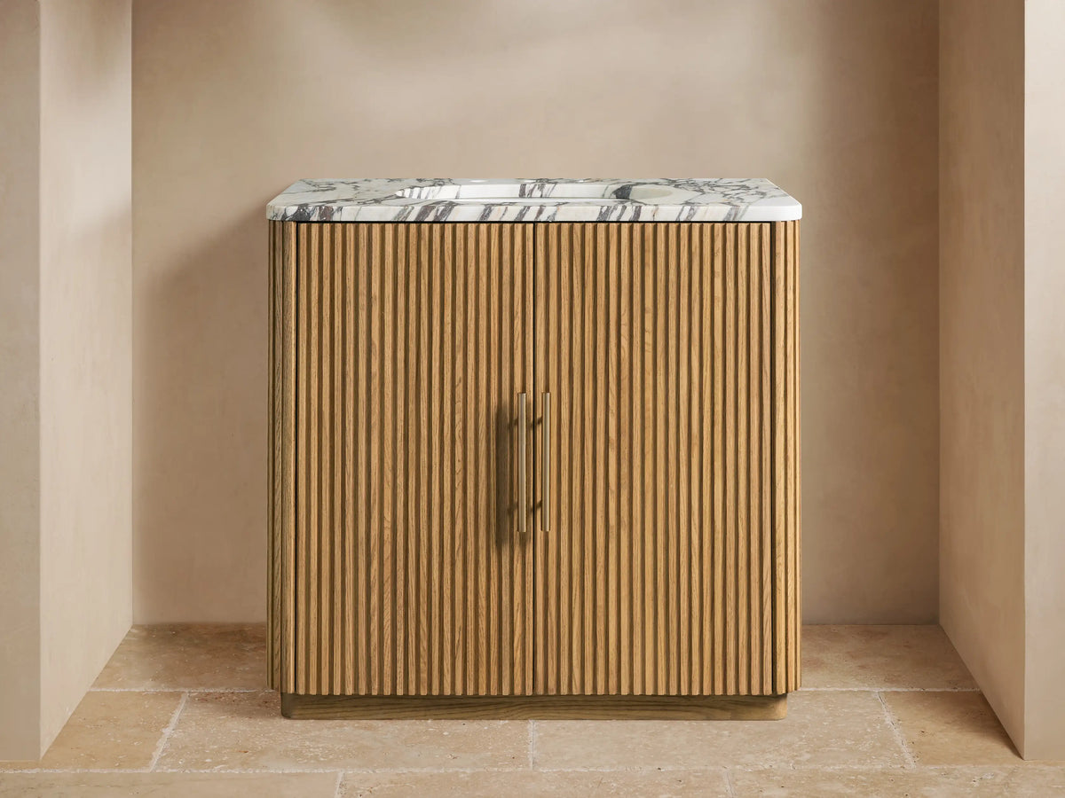Finnley Single Vanity: light oak wood with fluted doors, antique brass handles, and a white Carrara marble top with a sink.
