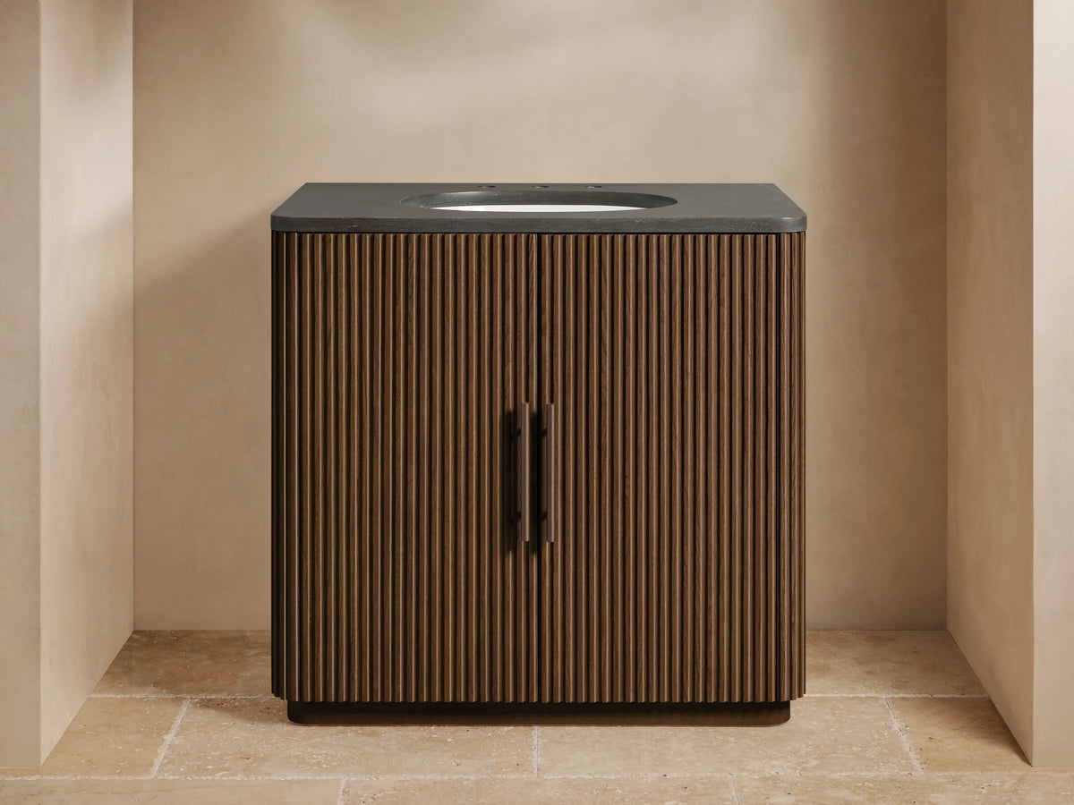 Finnley Single Vanity: dark wood fluted cabinet, bluestone top, white oval sink, bronze faucet, in a modern beige bathroom.