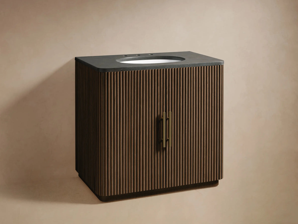 Finnley Single Vanity: dark brown fluted wood, floating. Dark gray stone top, white oval sink, brass faucet, light brown wall.