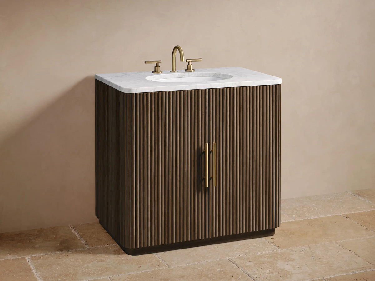 Finnley Single Vanity's open fluted dark oak door with gold handle. Inside: light wood shelf with rustic bowl and stacked beige towels.