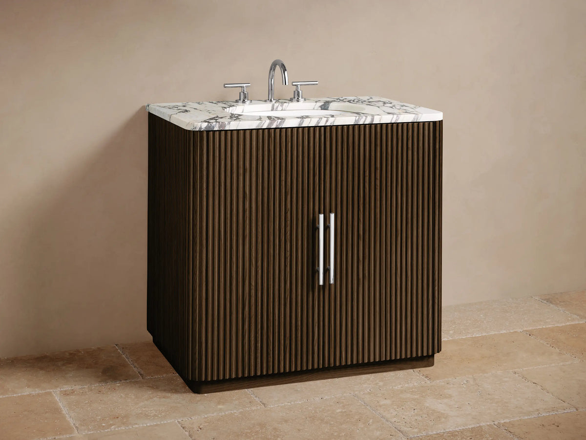 Finnley Single Vanity close-up: dark fluted wood door, white marble top. Open cabinet shows beige towels and toiletries.