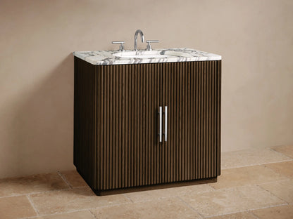 Finnley Single Vanity close-up: dark fluted wood door, white marble top. Open cabinet shows beige towels and toiletries.