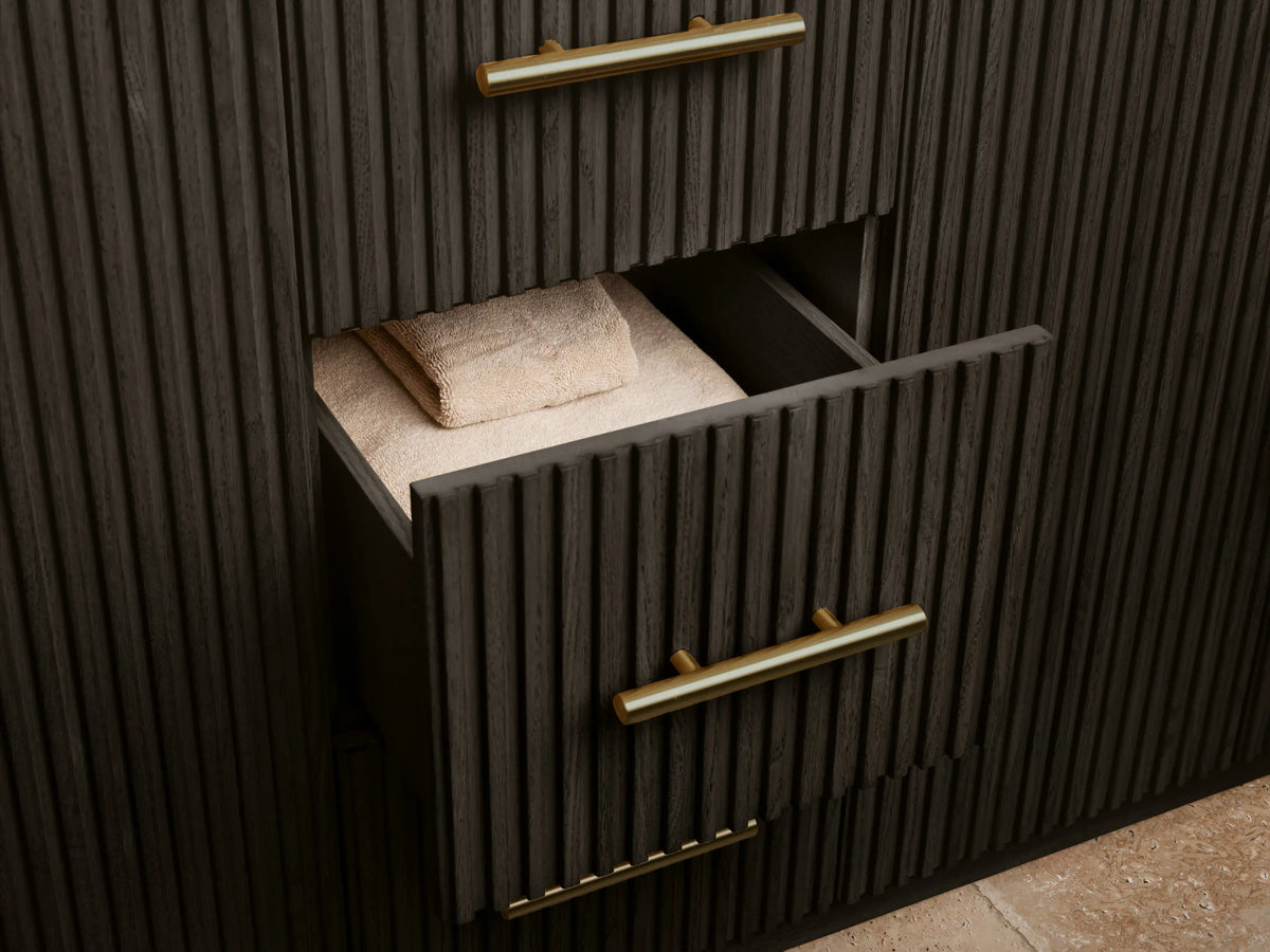 Finnley vanity's open dark wood cabinet door with ribbed detail & brass pull. Inside are shelves with stacked white towels & a sea sponge in a bowl.