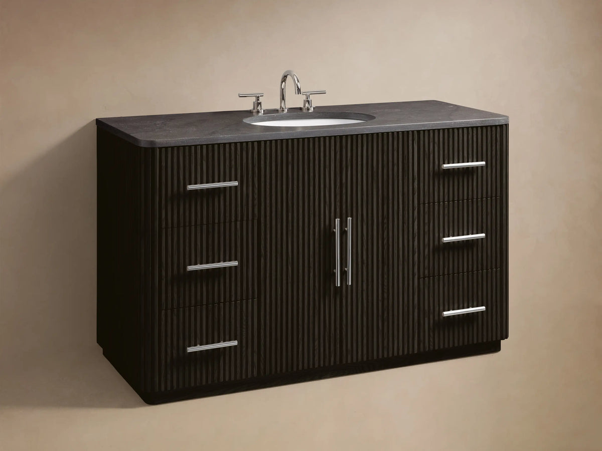 Close-up of Finnley Wide Single Vanity, dark fluted drawer with polished nickel pull partially open, showing towels & bottles. Dark stone top.