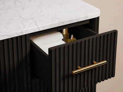 The image shows a close-up of the Finnley Single Wide Vanity with vertically grooved dark wood doors. Brass handles adorn the drawers and doors, which are slightly ajar, revealing folded white towels and a beige box on the inner shelves.