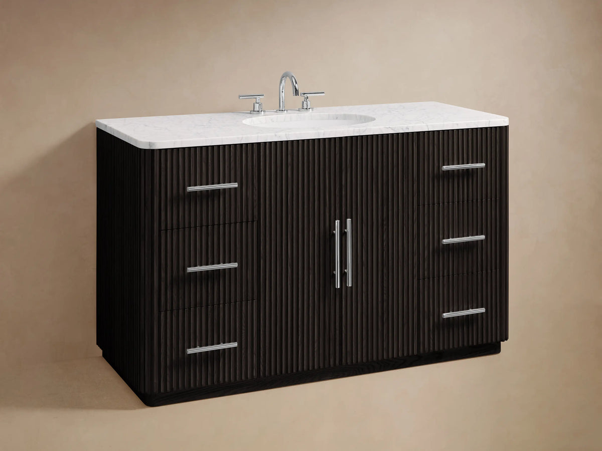 The Finnley Single Wide Vanity features a dark oak wood drawer with horizontal grooves, a silver handle, and a marble top. Inside are a folded towel and toiletries.