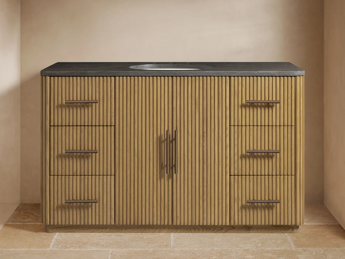 Finnley Wide Single Vanity: fluted light wood cabinet, dark stone top with oval sink, bronze faucet, and hardware in a bathroom.