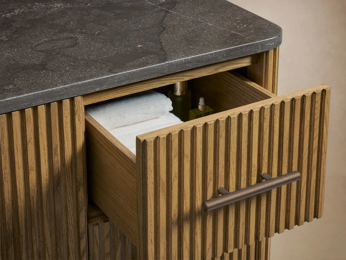 Close-up of Finnley Wide Single Vanity's ribbed oak door slightly open, revealing shelves with white towels, next to ribbed drawers with antique bronze handles.