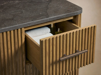 Close-up of Finnley Wide Single Vanity's ribbed oak door slightly open, revealing shelves with white towels, next to ribbed drawers with antique bronze handles.