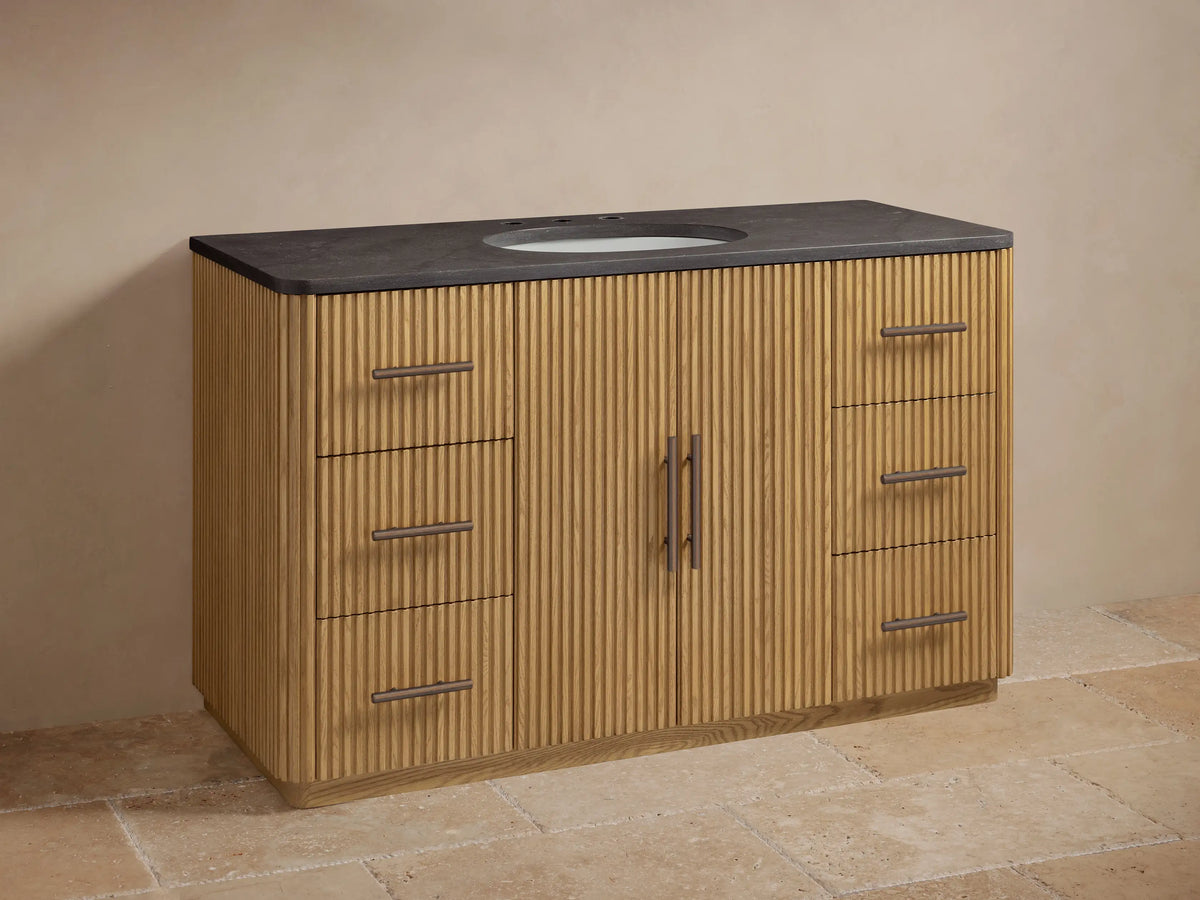 Finnley Wide Single Vanity: Fluted light oak cabinet, dark stone top, white oval sink, bronze faucet. 6 drawers, 2 doors.
