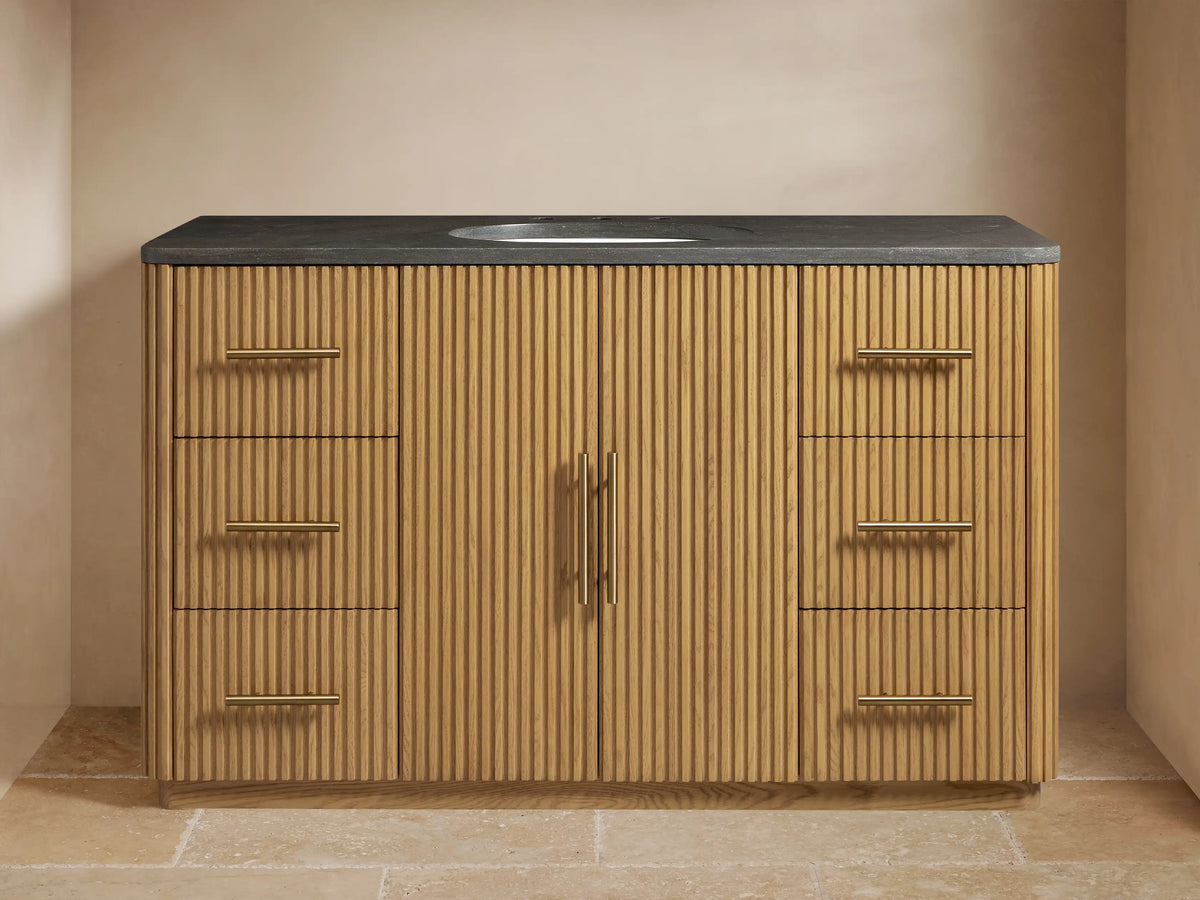 Finnley Single Wide Vanity with vertical oak slats, black stone top, oval sink, brass hardware, dual faucet setup.