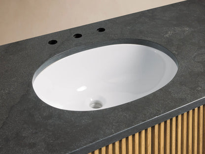 Oval ceramic undermount sink set in dark stone vanity top with polished nickel faucet and soap dish. Fluted oak wood detail visible below.
