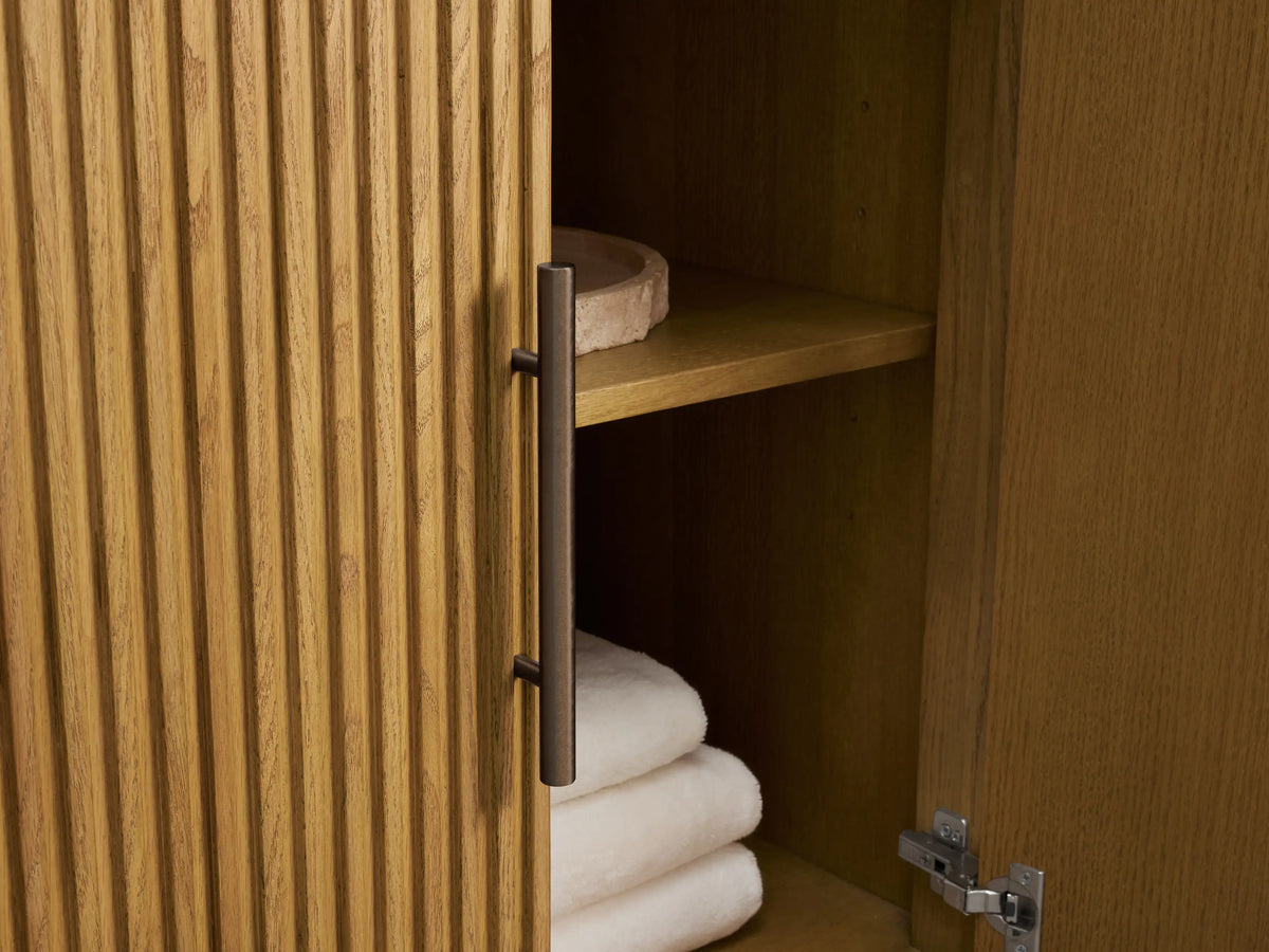 Close-up of Finnley Wide Single Vanity. Light wood fluted cabinet, white marble top, bronze pulls. Open drawer with towel, bottles.