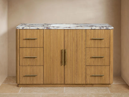 Finnley Wide Single Vanity. Natural oak ribbed cabinet with gold hardware, white marble top, oval sink, in a beige room setting.