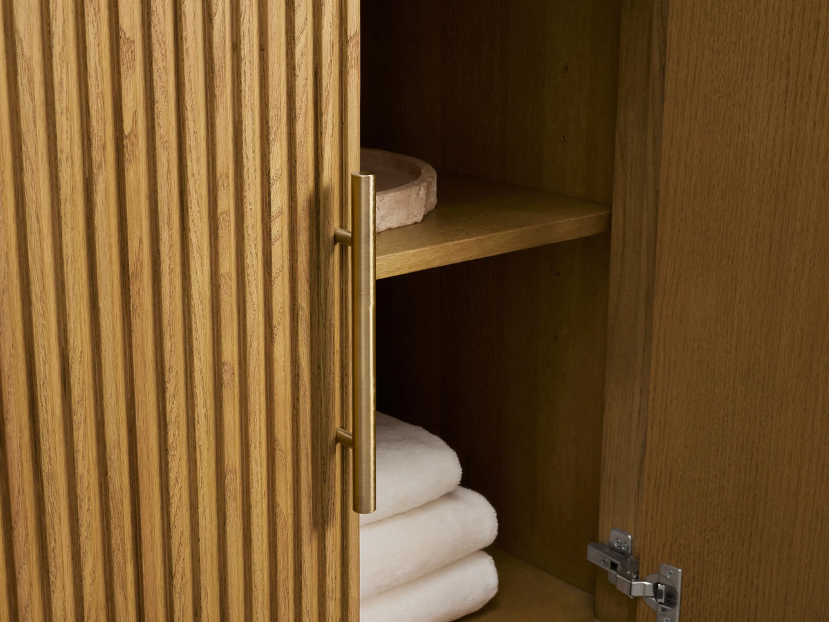 Close-up of Finnley Wide Single Vanity. Fluted light oak cabinet, Carrara marble top, brass handles. Drawer open, showing towel and bottles.