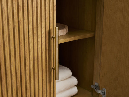 Close-up of Finnley Wide Single Vanity. Fluted light oak cabinet, Carrara marble top, brass handles. Drawer open, showing towel and bottles.
