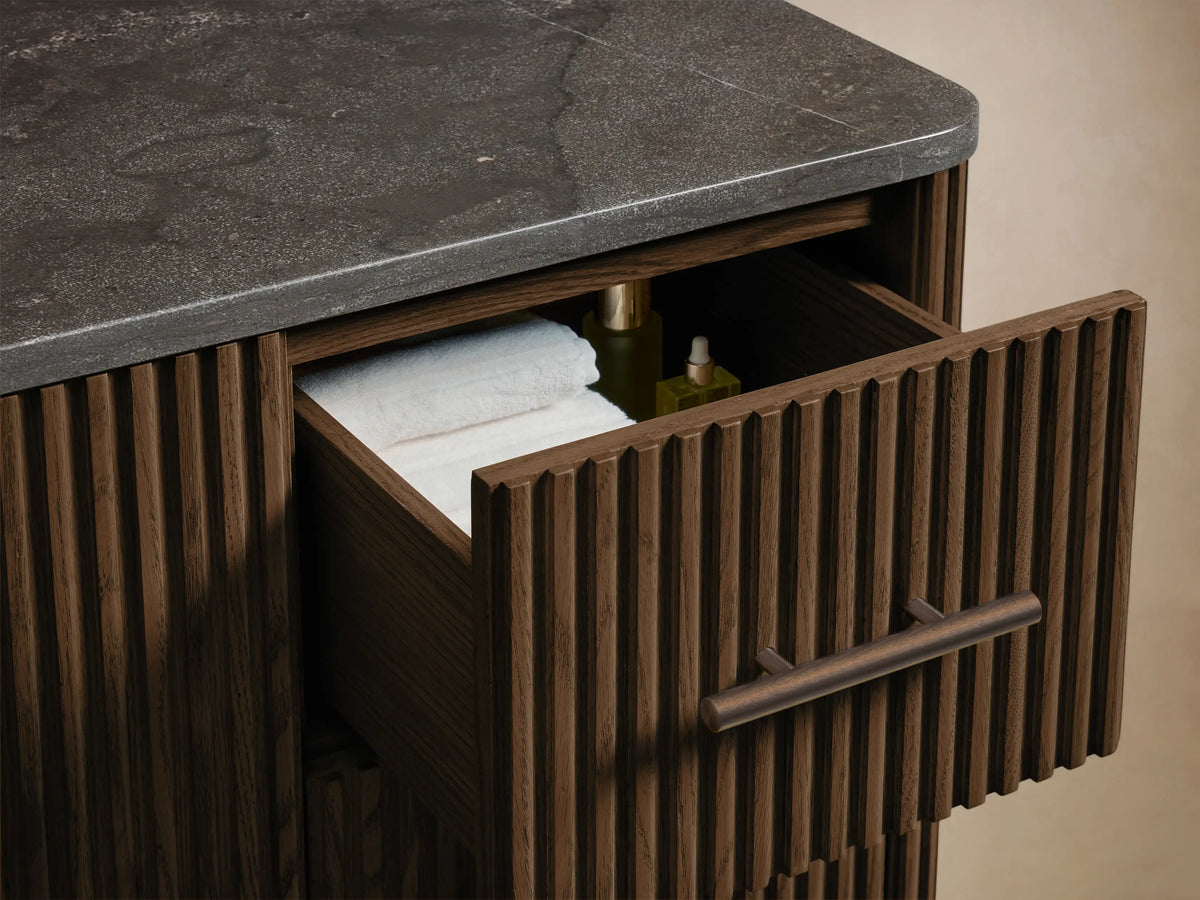 Close-up of Finnley Wide Single Vanity, dark oak wood with ribbed texture & antique bronze handles. Open door shows shelf with box, towels.