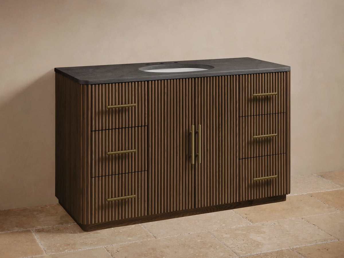 Finnley Wide Single Vanity: Dark brown wood with fluted panels, dark stone top, white oval sink, gold faucet & hardware.