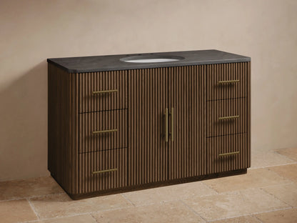 Finnley Wide Single Vanity: Dark brown wood with fluted panels, dark stone top, white oval sink, gold faucet & hardware.