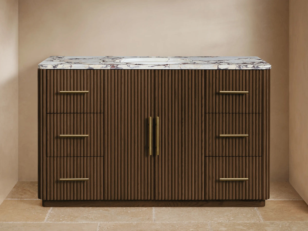 Finnley Wide Single Vanity: Dark brown fluted wood vanity with white marble top, gold widespread faucet, drawers, and cabinet doors.