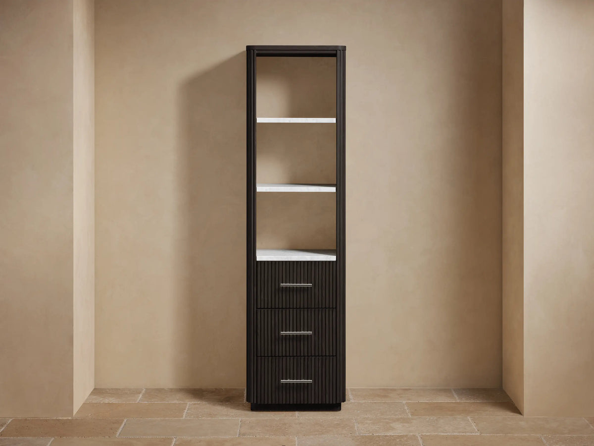 Tall dark brown Finnley bath cabinet with open shelves displaying decor/towels & three fluted drawers with silver pulls, against a beige wall.